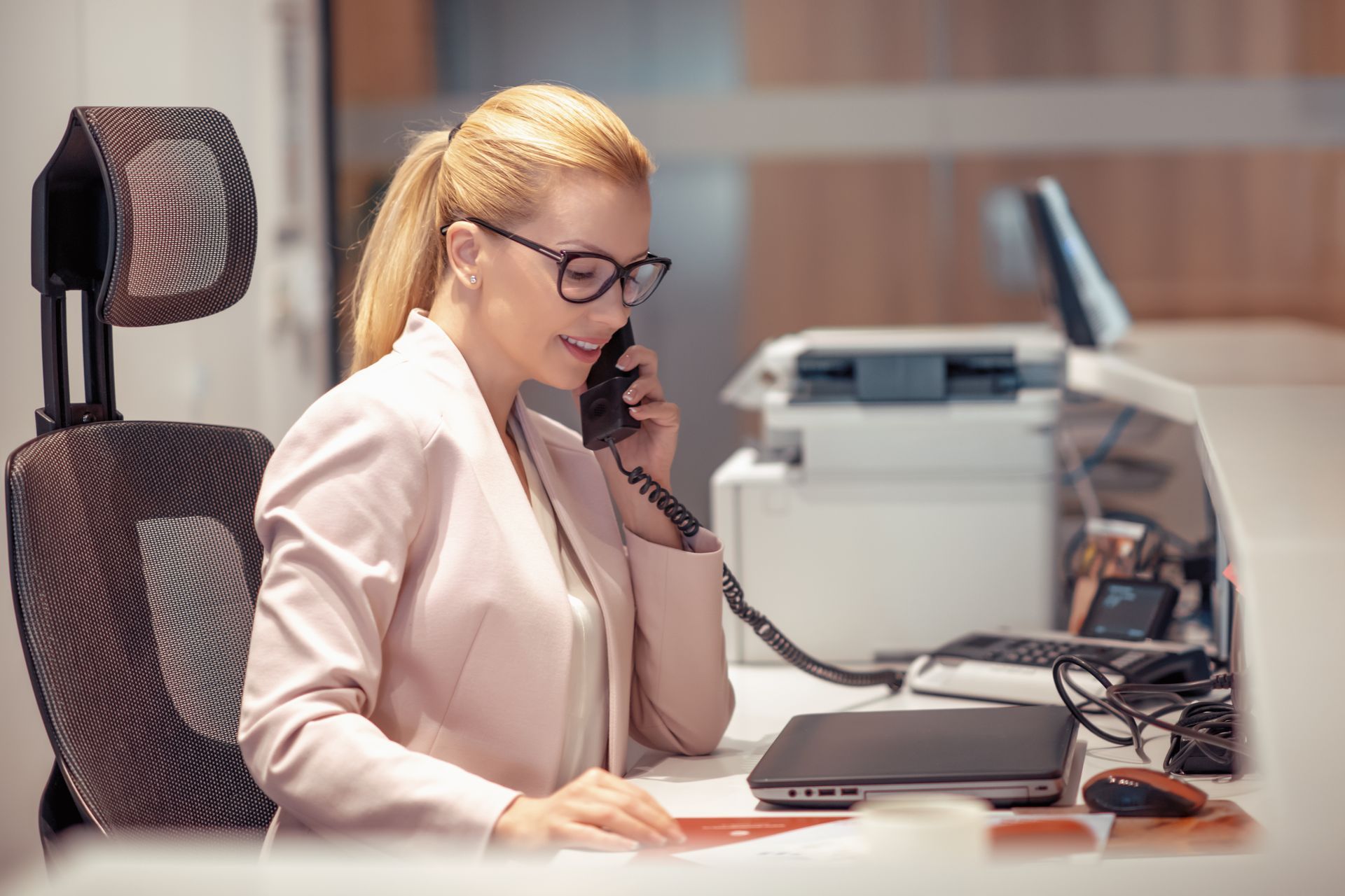 Une femme portant des lunettes parle au téléphone, à son bureau, avec un ordinateur et une imprimante à proximité.