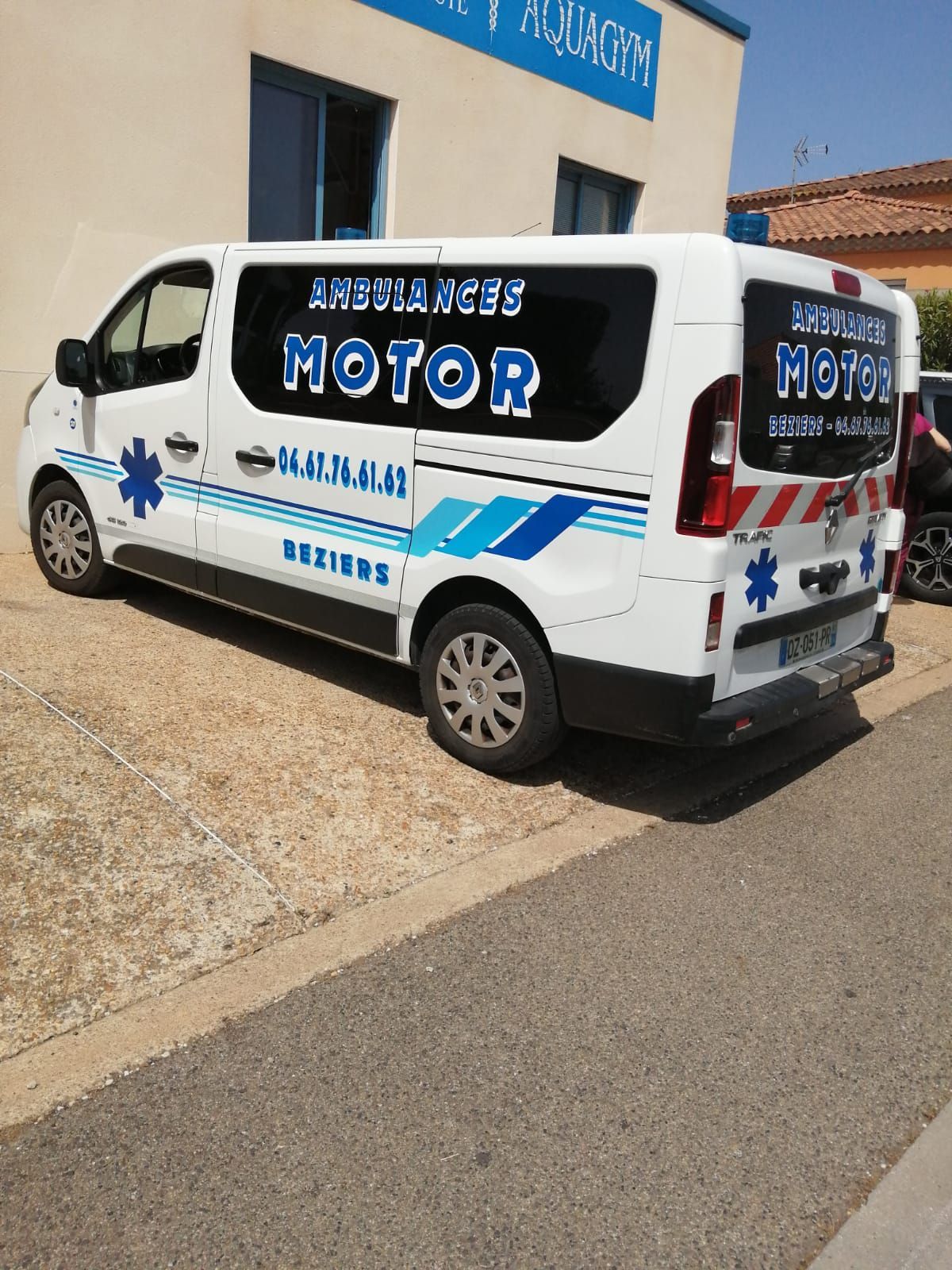 Une camionnette de dépannage blanche avec des autocollants bleus est garée à côté d'un bâtiment en plein soleil.