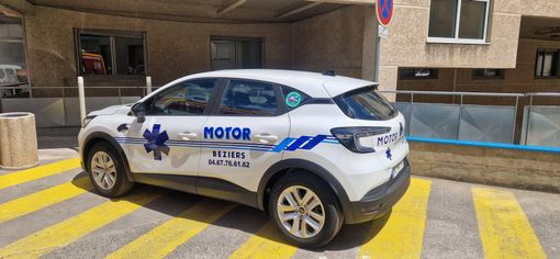 Un SUV électrique MOTOR blanc et bleu est garé dans une rue à côté de bâtiments.