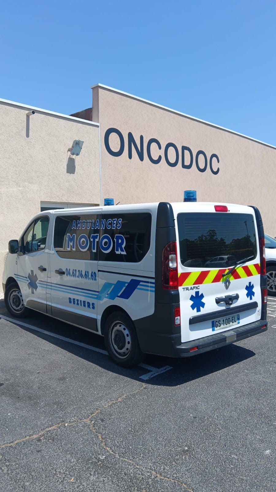 Une ambulance blanche avec des marquages ​​bleus et rouges est garée devant un bâtiment de l'ONCODOC sous un ciel dégagé.
