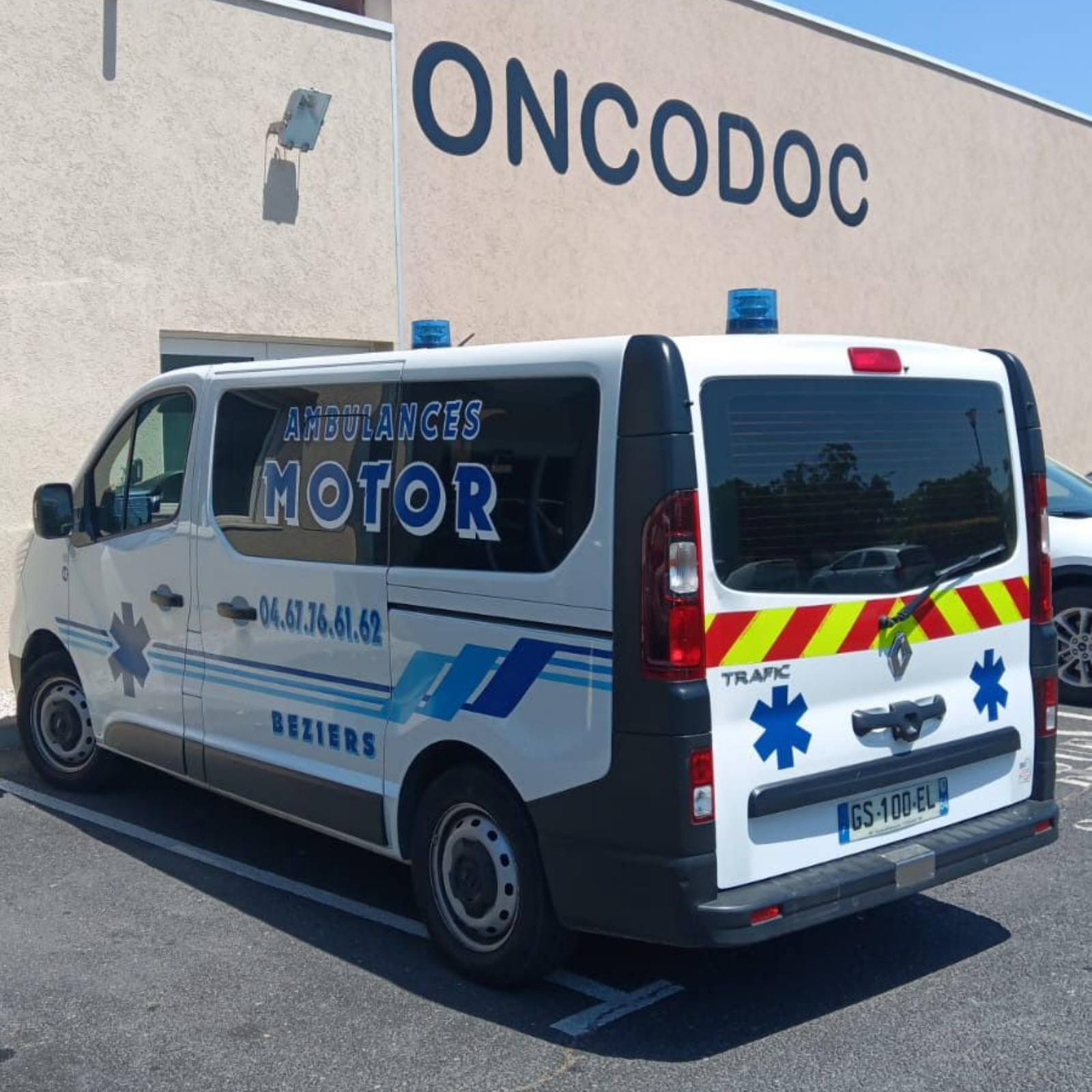 Fourgonnette d'urgence blanche avec gyrophares bleus stationnée devant le bâtiment ONCODOC, vue arrière avec bandes réfléchissantes rouges et jaunes.