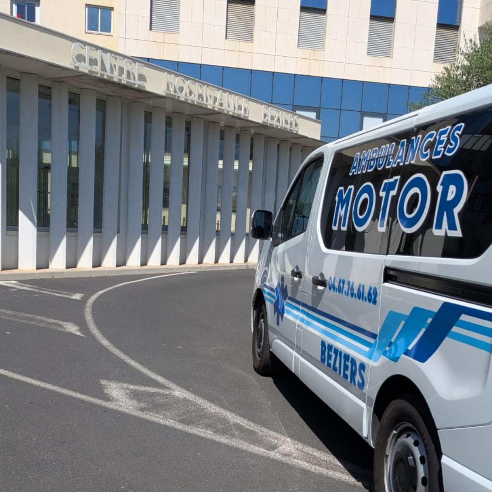 Une ambulance blanche avec des inscriptions bleues passe devant un bâtiment hospitalier à colonnes blanches.
