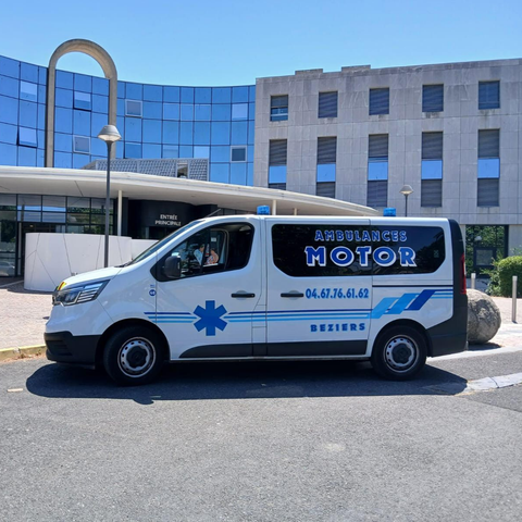Un véhicule blanc de transport médical est garé devant un immeuble bleu moderne dans une rue ensoleillée.
