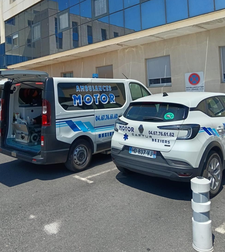 Une ambulance et un VSL stationnés devant un bâtiment en verre bleu, un des véhicules a les portes arrière ouvertes.