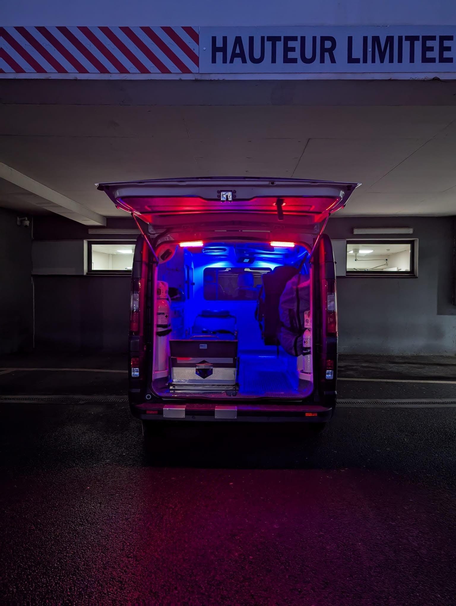 Ambulance avec portes arrière ouvertes, gyrophares bleus et rouges clignotant sous un panneau de hauteur réduite, la nuit