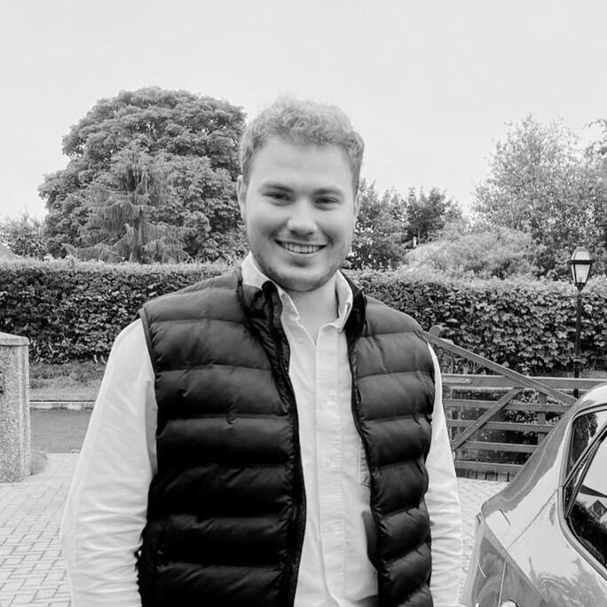 Man in a black vest and white shirt smiles outside. Bushes and a car are in the background.