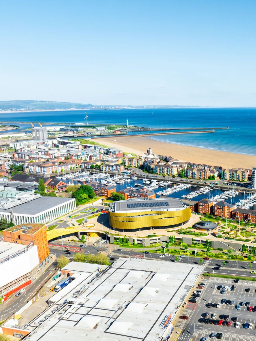Coastal cityscape with beach, marina, gold-colored building, and blue sea under a clear sky.