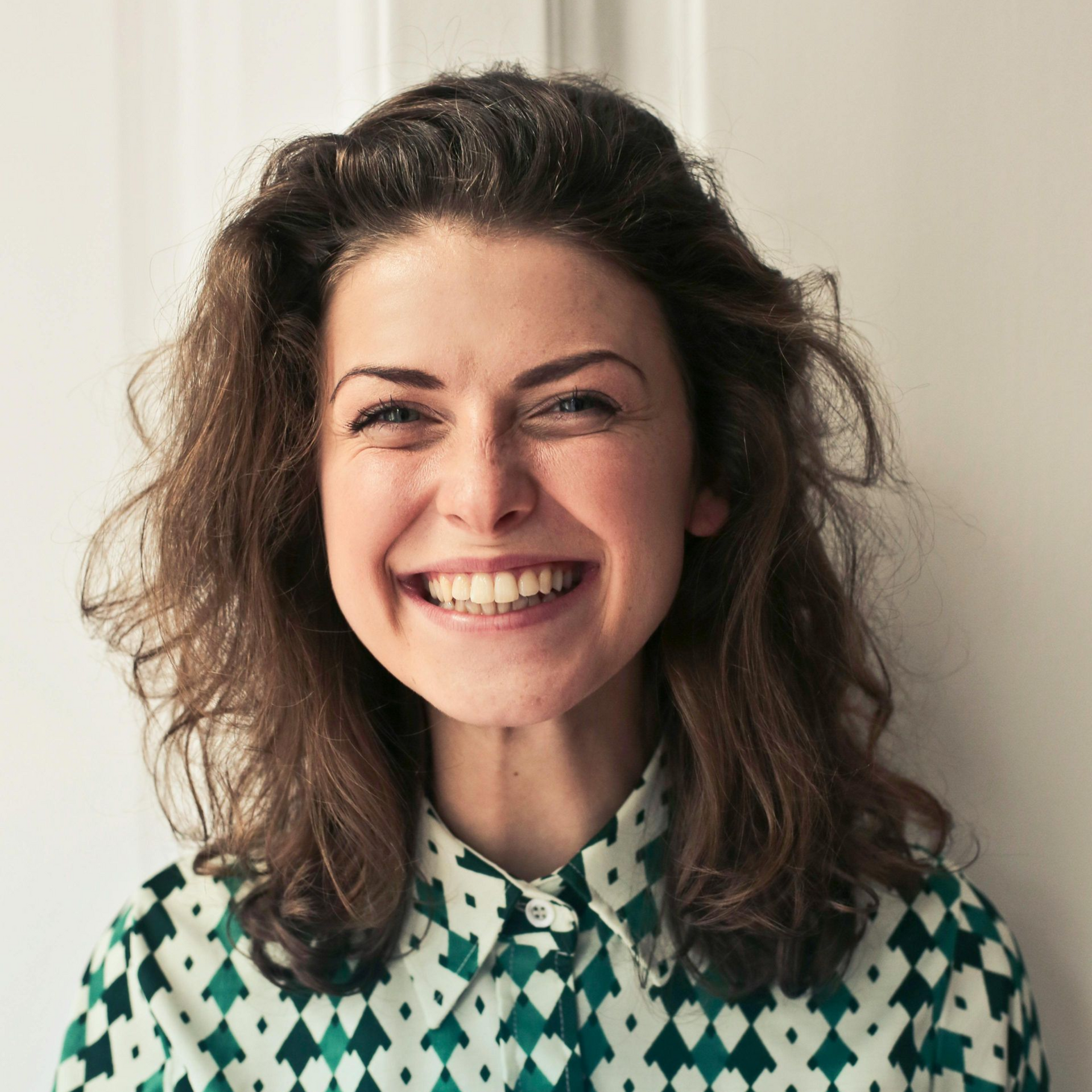 Woman with brown hair smiles broadly, wearing a patterned green and white collared shirt, against a white wall.