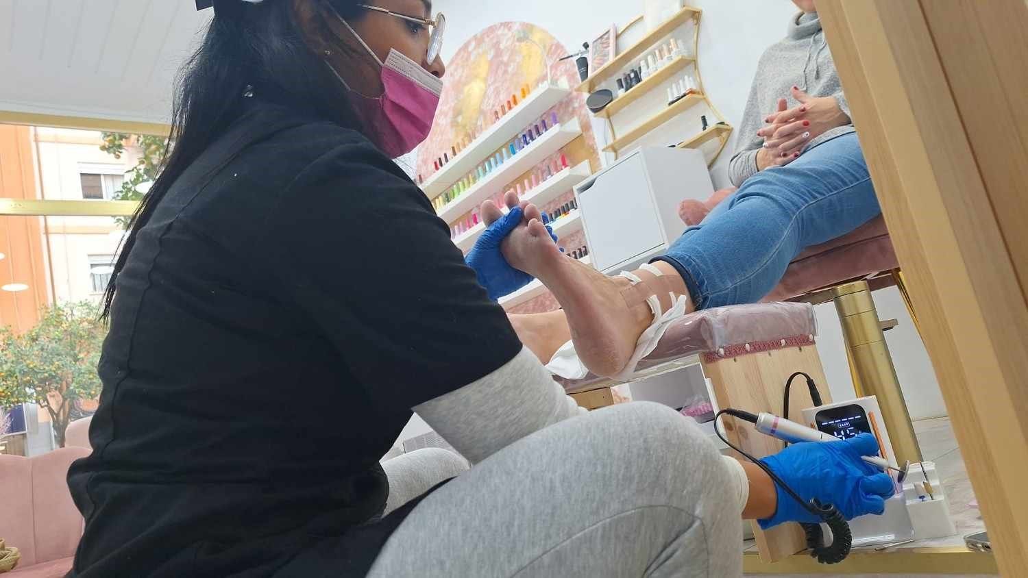Una persona pinta las uñas de los pies de otra persona en un salón de belleza, con estantes llenos de esmaltes.