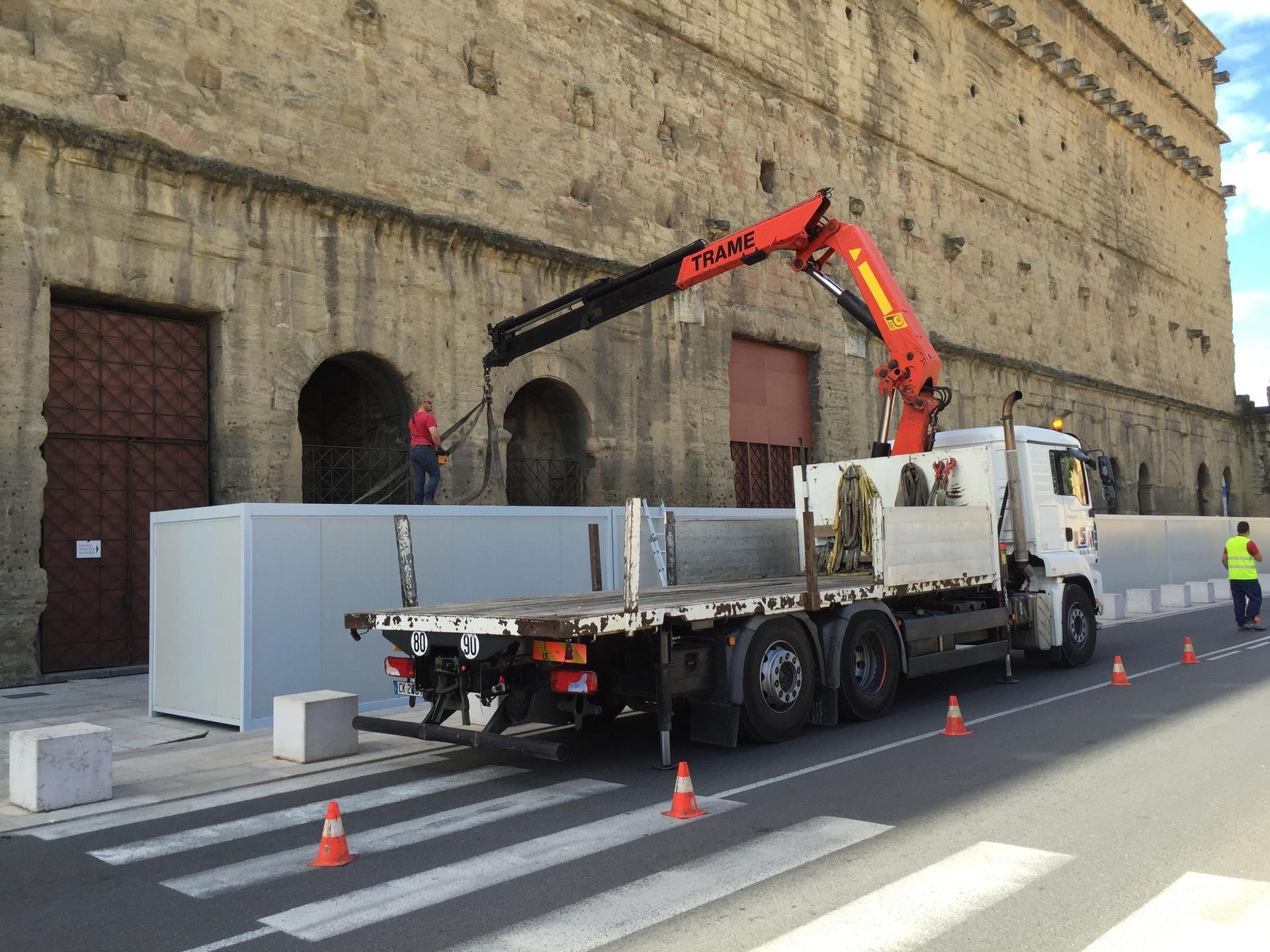 Camion-grue déchargeant des barrières grises à côté d'un bâtiment en pierre dans une rue de la ville, avec des cônes et un ouvrier à proximité.