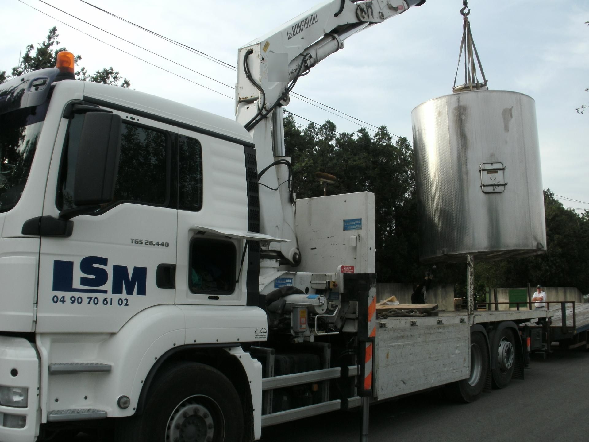 Camion-grue blanc soulevant un grand réservoir argenté sur un chantier.