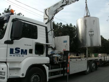 Camion-grue blanc soulevant un grand réservoir argenté sur un chantier.