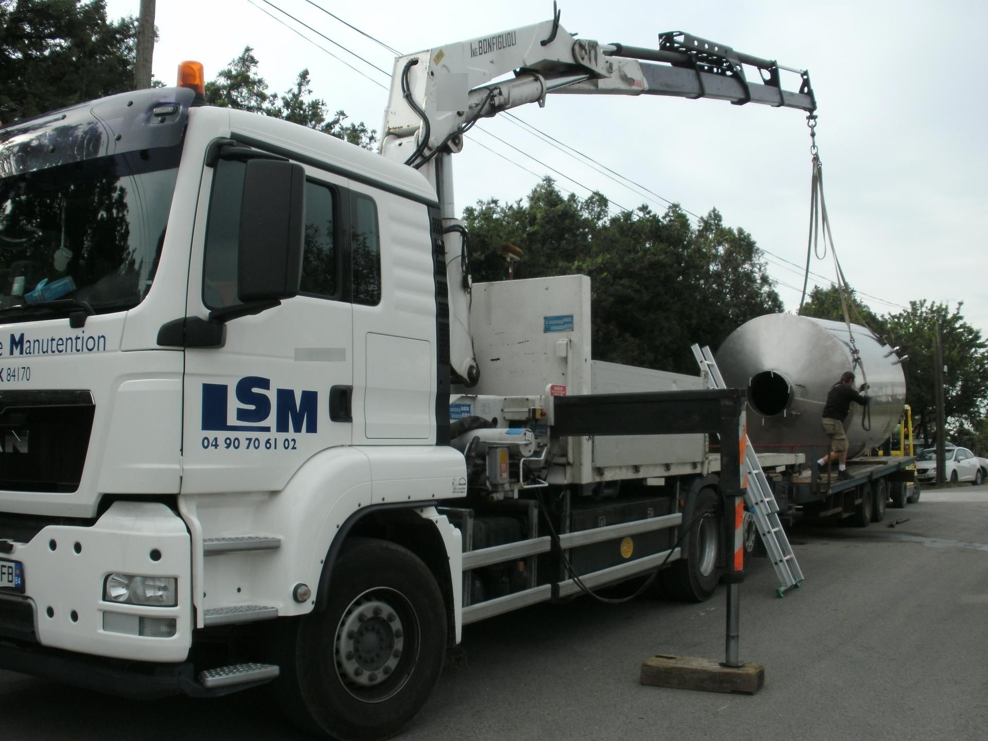 Camion malaxeur à béton blanc LSM avec bras articulé, stationné dans une rue à côté d'une échelle et de cônes de signalisation.