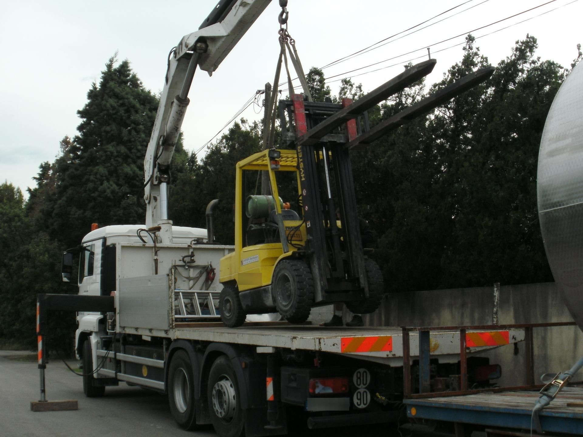 Un chariot élévateur jaune sur un camion plateau est soulevé par une grue à l'extérieur.