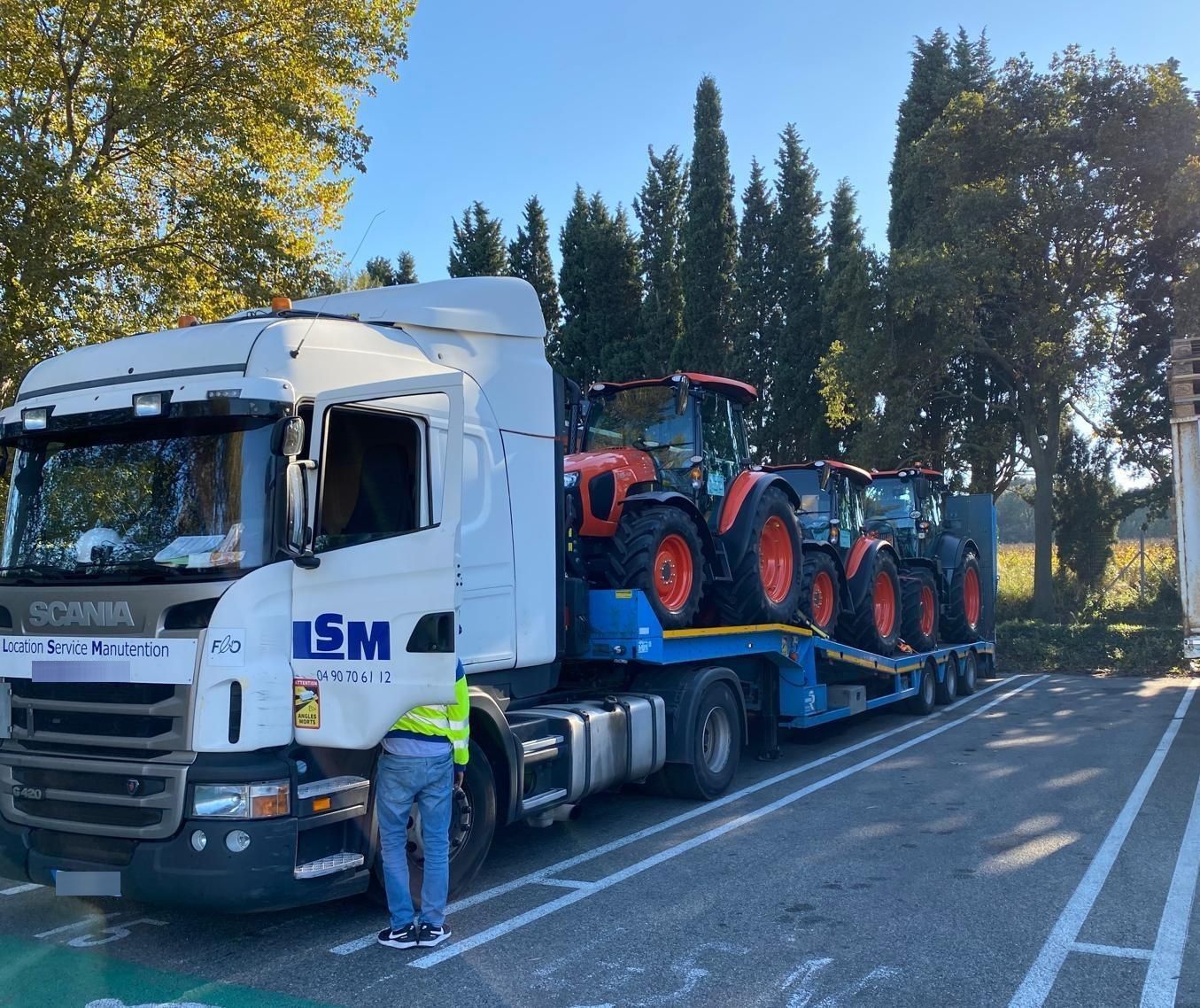 Un camion blanc transporte des tracteurs rouges sur une remorque à plateau, un ouvrier se tenant à côté, au bord de la route.