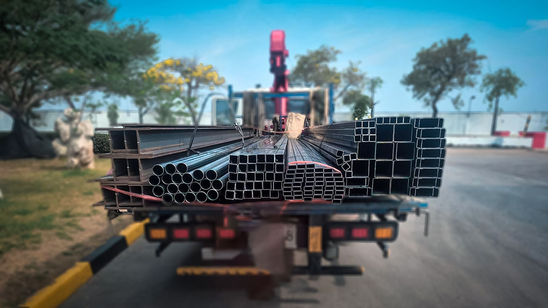 Camion chargé de tuyaux métalliques noirs empilés en bord de route, avec une grue rouge et des arbres en arrière-plan.