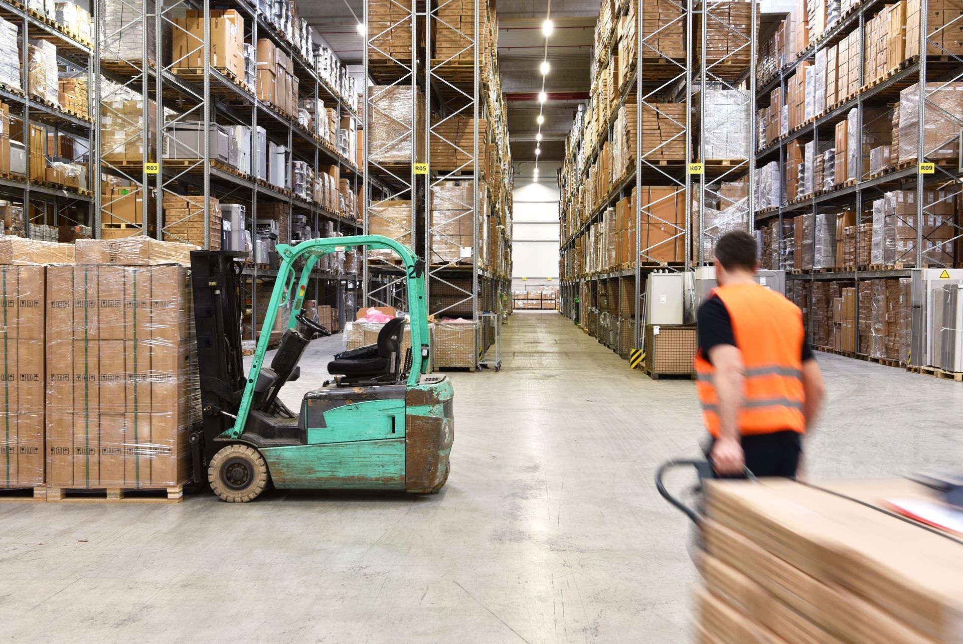Allée d'entrepôt avec un chariot élévateur, des cartons empilés et un employé portant un gilet de sécurité orange.