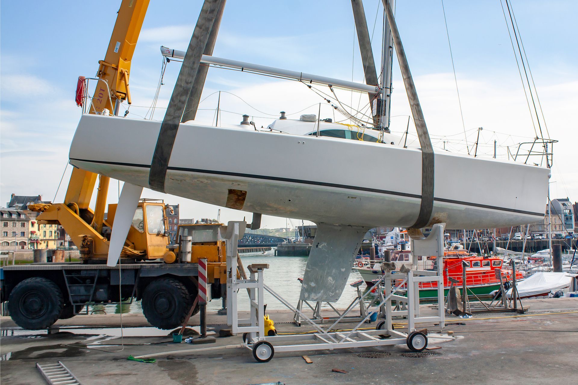 La coque d'un grand voilier blanc est soulevée par une grue dans un chantier naval, avec des ouvriers et du matériel à proximité.