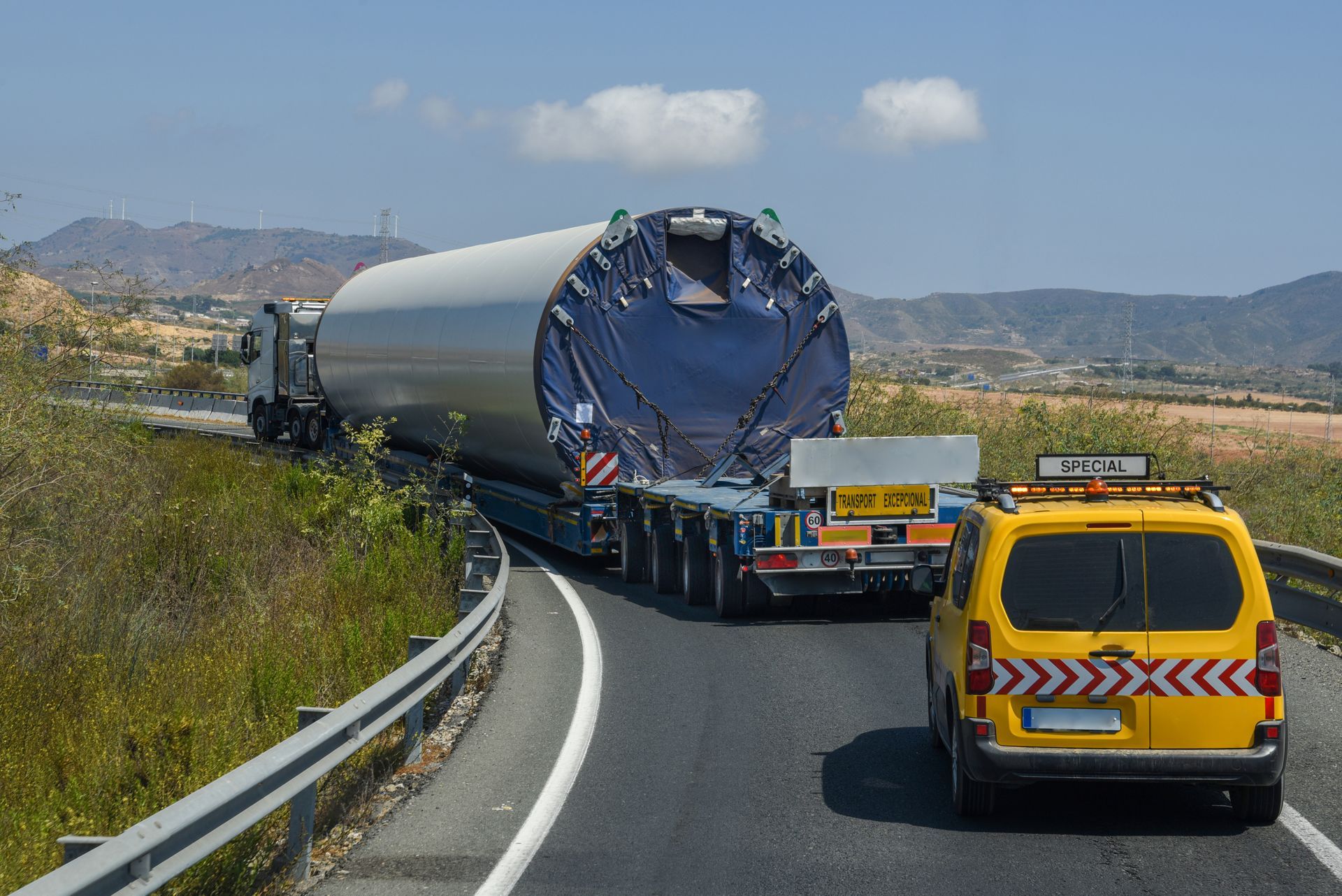 Un camion transportant une énorme charge cylindrique argentée circule sur une route sinueuse, suivi d'un véhicule d'escorte jaune.