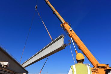 Une grue soulève une grosse poutre d'acier sur un chantier de construction sous un ciel bleu dégagé.