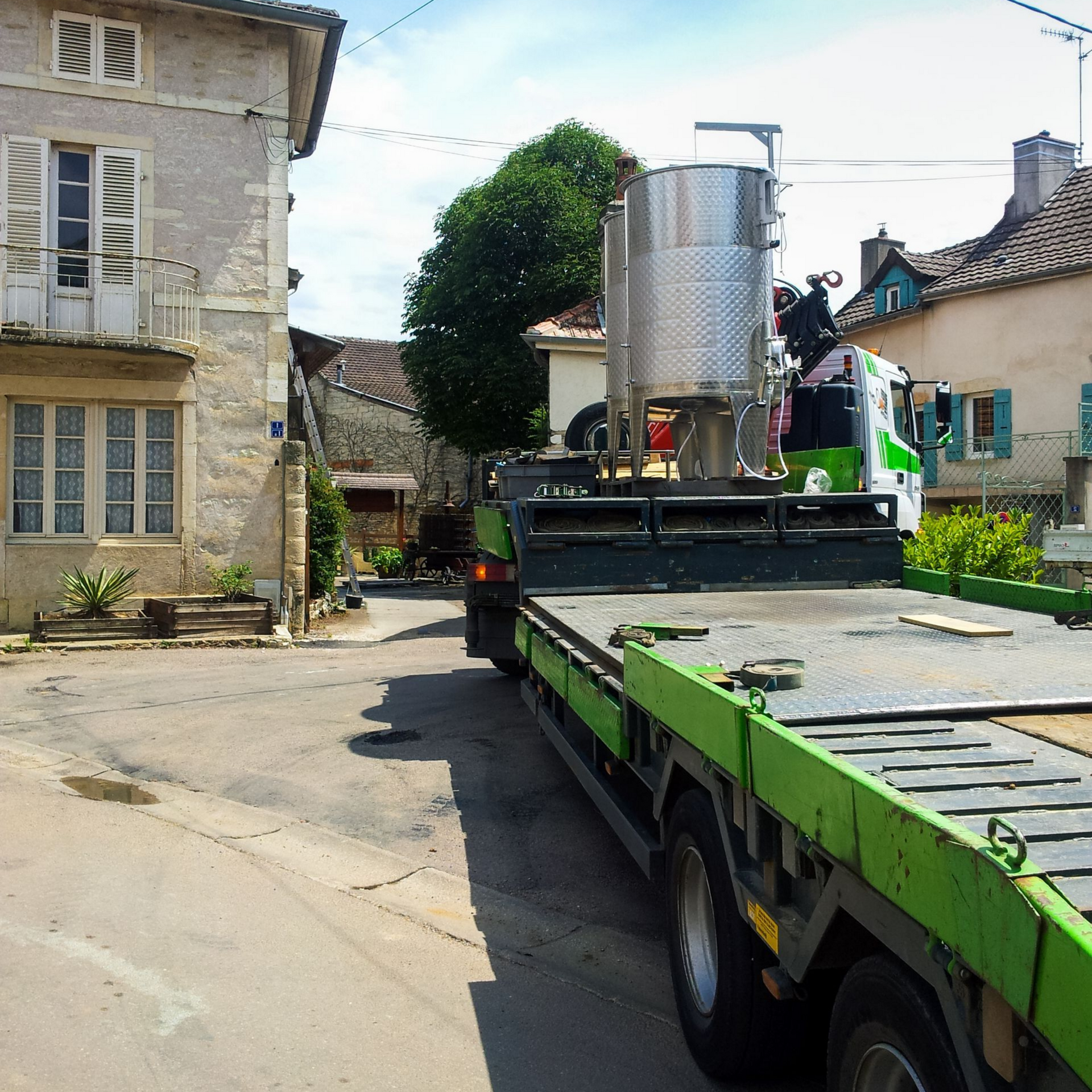 Un camion-plateau vert transportant une cuve en acier circule dans une rue étroite entre de vieux bâtiments en pierre.