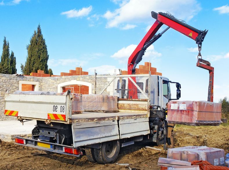 Camion de chantier déchargeant des briques à l'aide d'une grue rouge sur un chantier de construction.