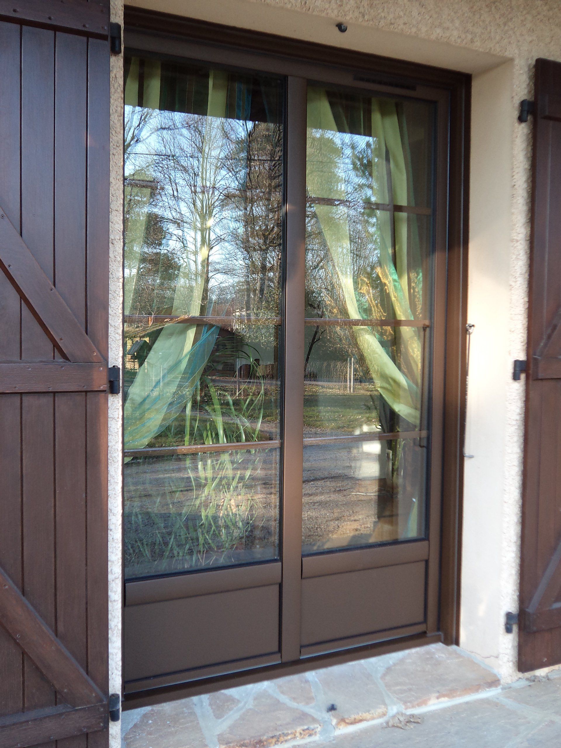 Porte double marron avec panneaux de verre et volets assortis ; rideaux verts à l'intérieur.