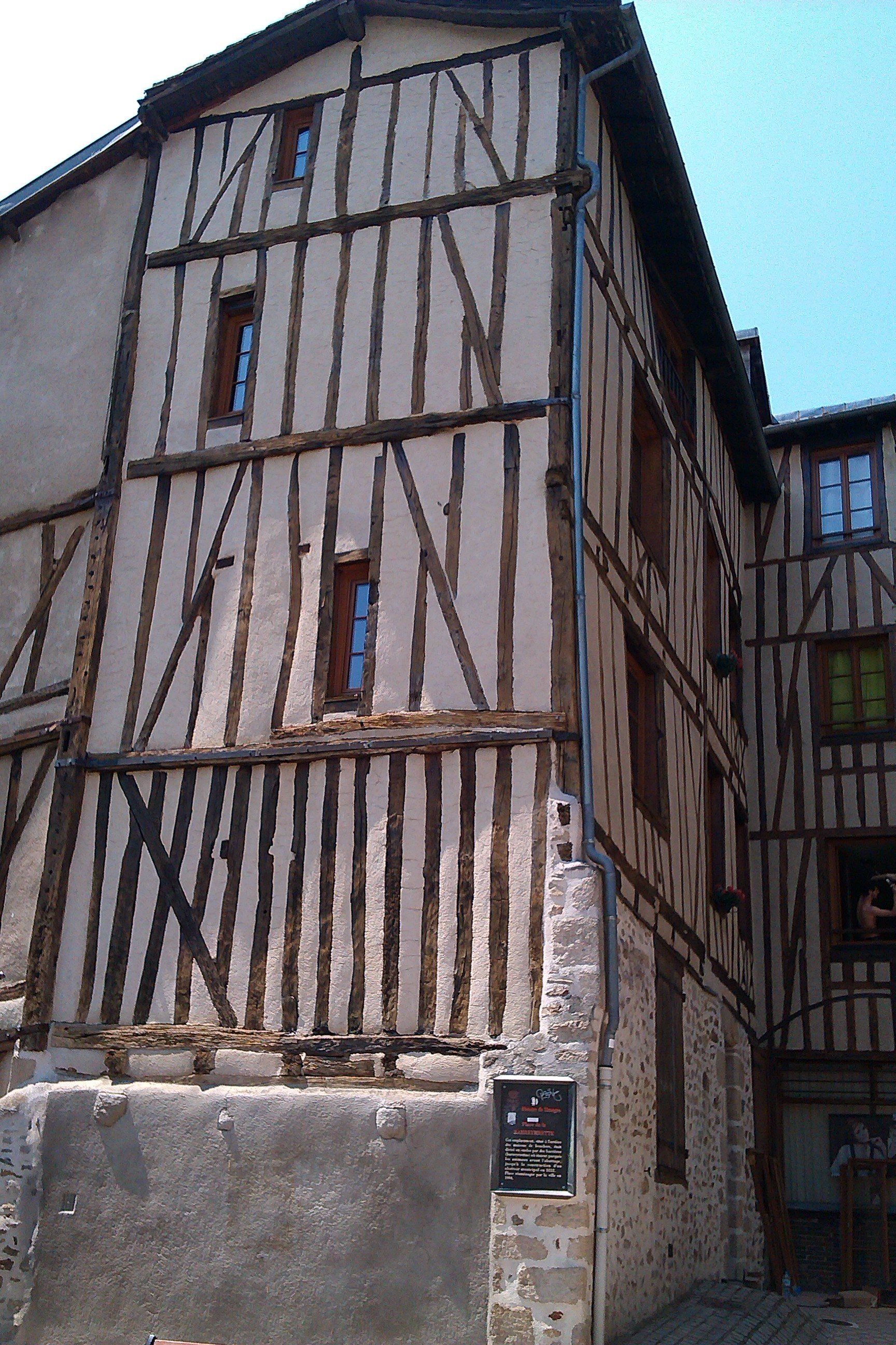 Bâtiment à colombages situé à l'angle d'une rue étroite. Façade beige et marron, petites fenêtres.