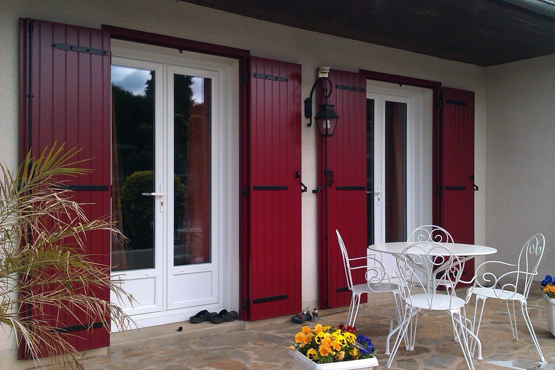 Des volets rouges encadrent des portes blanches donnant sur une terrasse meublée de mobilier de jardin blanc et agrémentée de fleurs en pot.