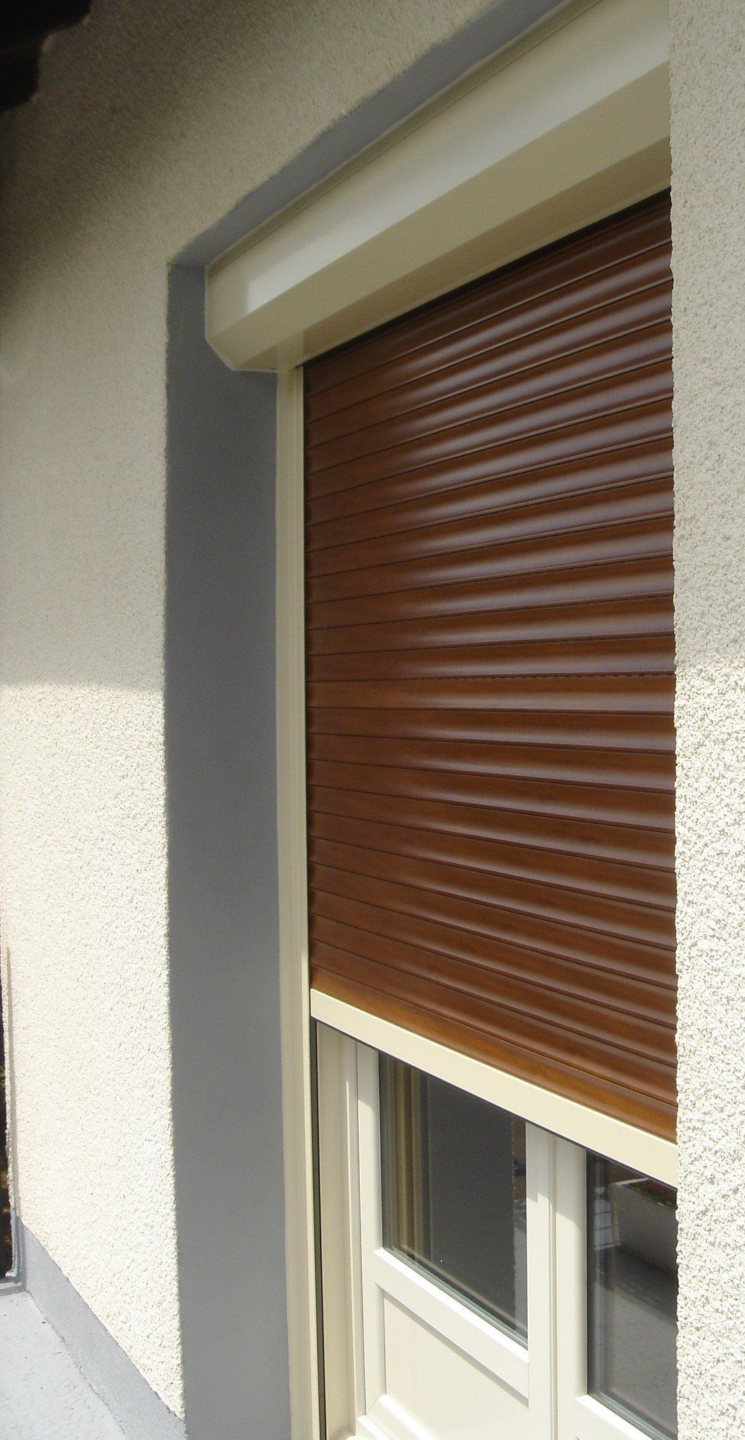 Volet roulant extérieur marron recouvrant partiellement une fenêtre. Boiseries beiges, mur en stuc.