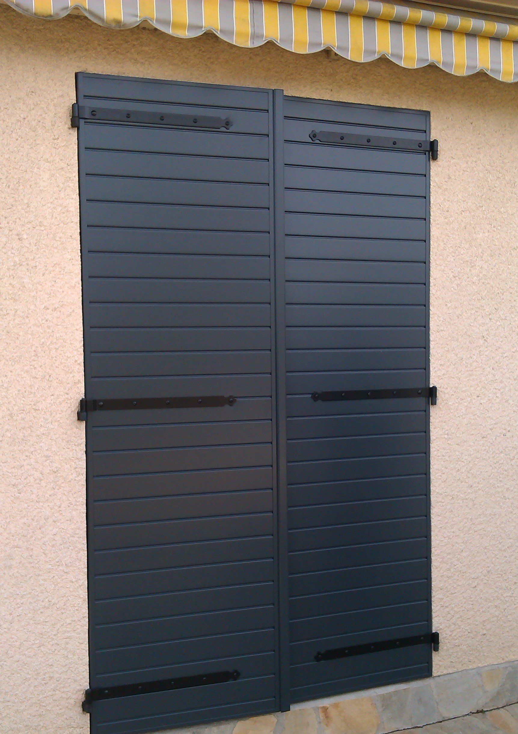 Volets noirs sur un bâtiment beige avec un auvent rayé en arrière-plan.