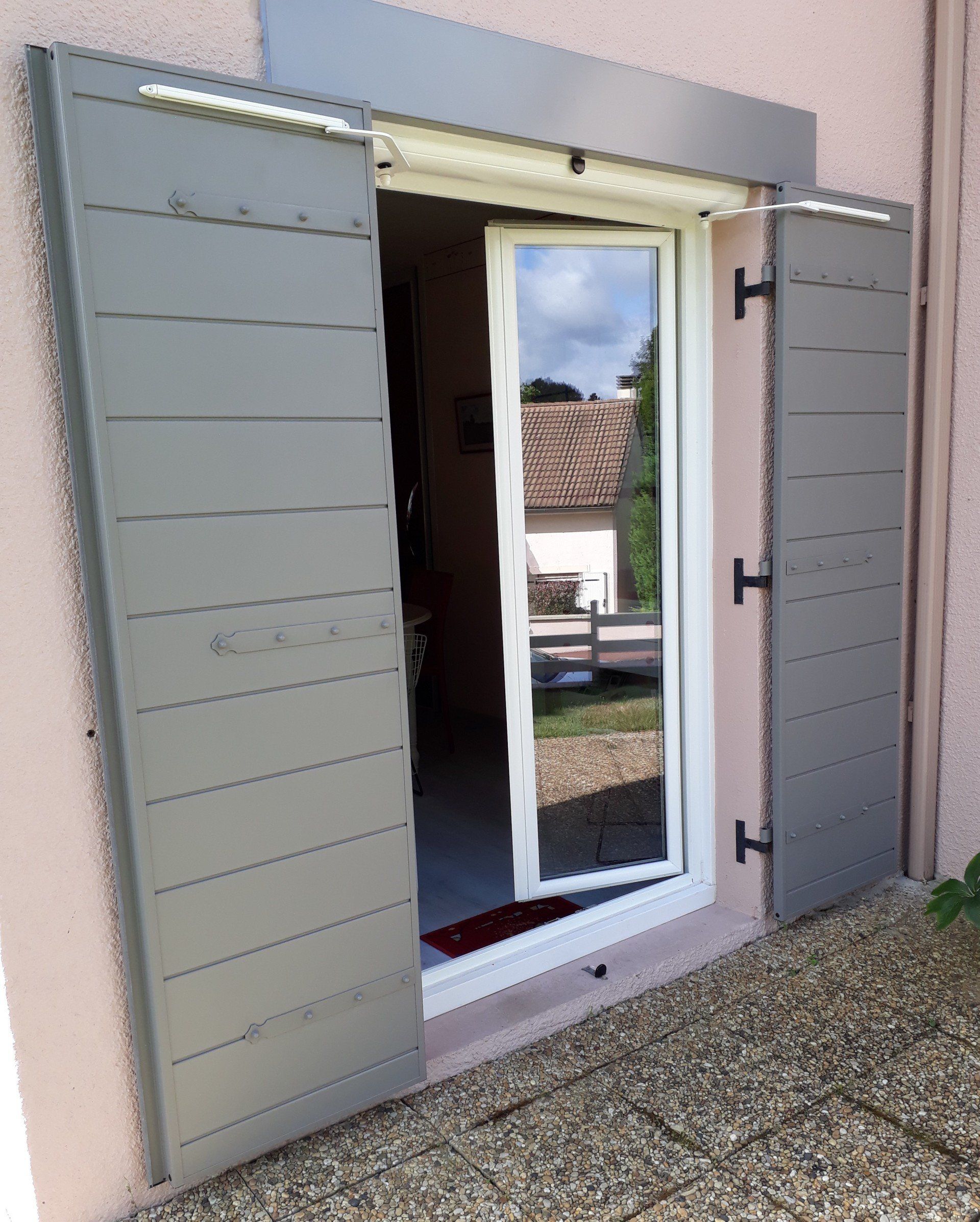 Des volets gris encadrent une porte vitrée à cadre blanc, partiellement ouverte, encastrée dans un mur en stuc rose.