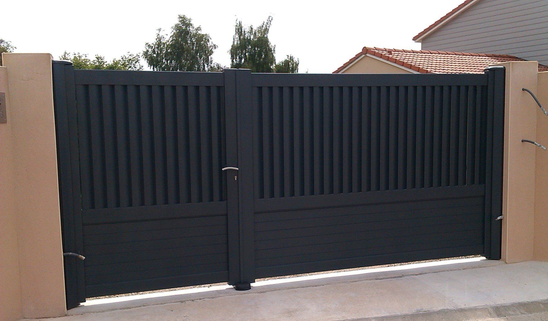 Portail double en métal gris foncé devant une maison. Barreaux verticaux, piliers beige clair.