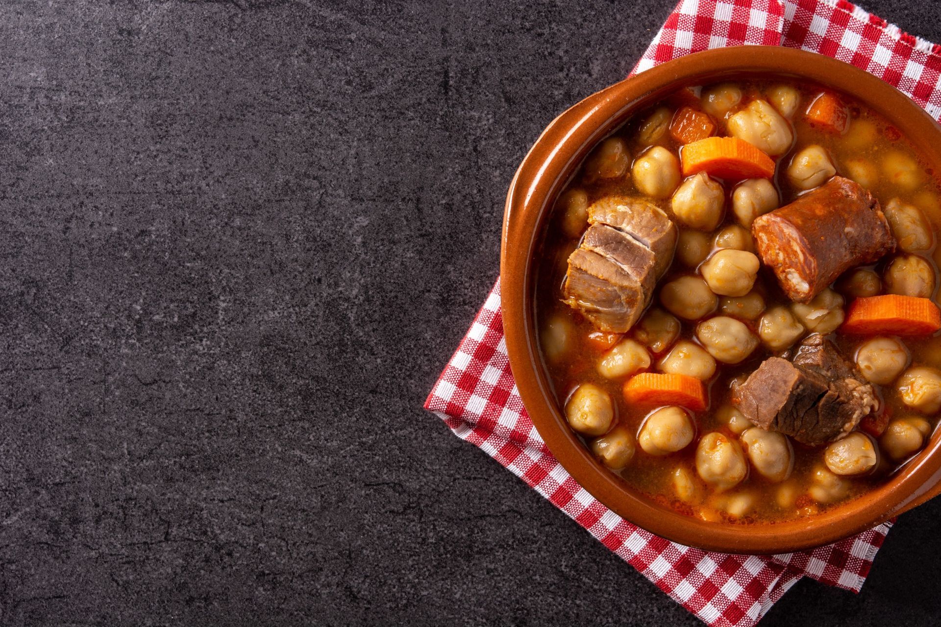 Un tazón de sopa con garbanzos, zanahorias y carne sobre una servilleta a cuadros sobre una mesa.