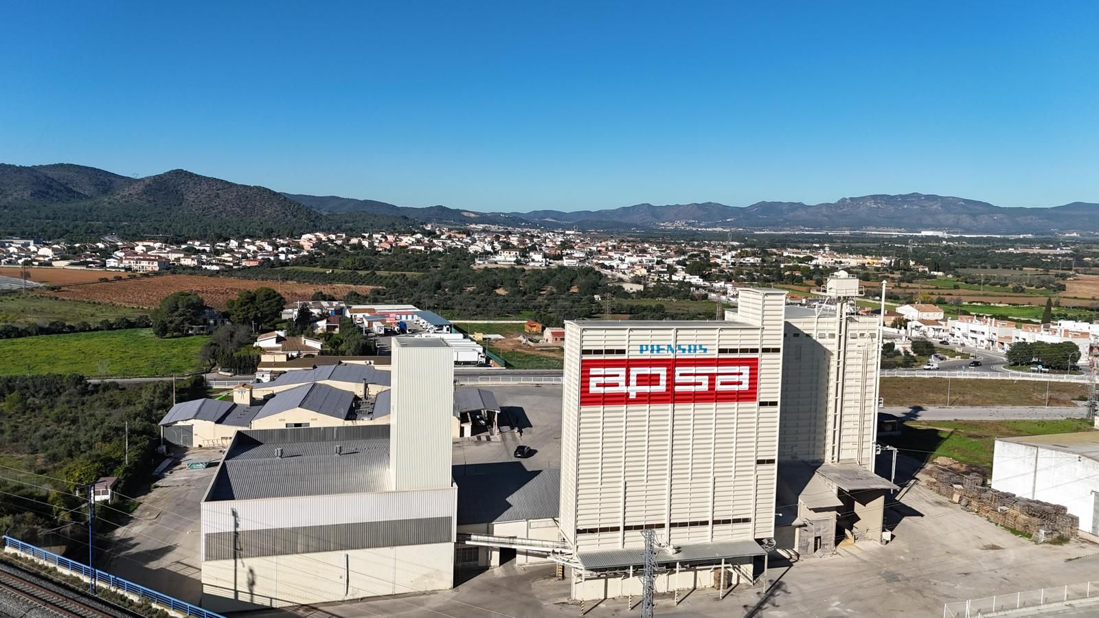 Vista aérea de la fábrica de APSA con un gran logotipo rojo, edificios y la ciudad al fondo bajo un cielo azul.