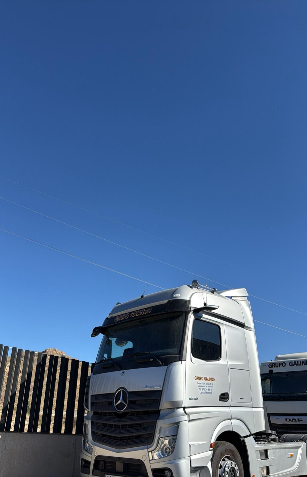 Camión semirremolque blanco con remolque estacionado bajo un cielo azul claro. El camión tiene detalles en verde y rojo.