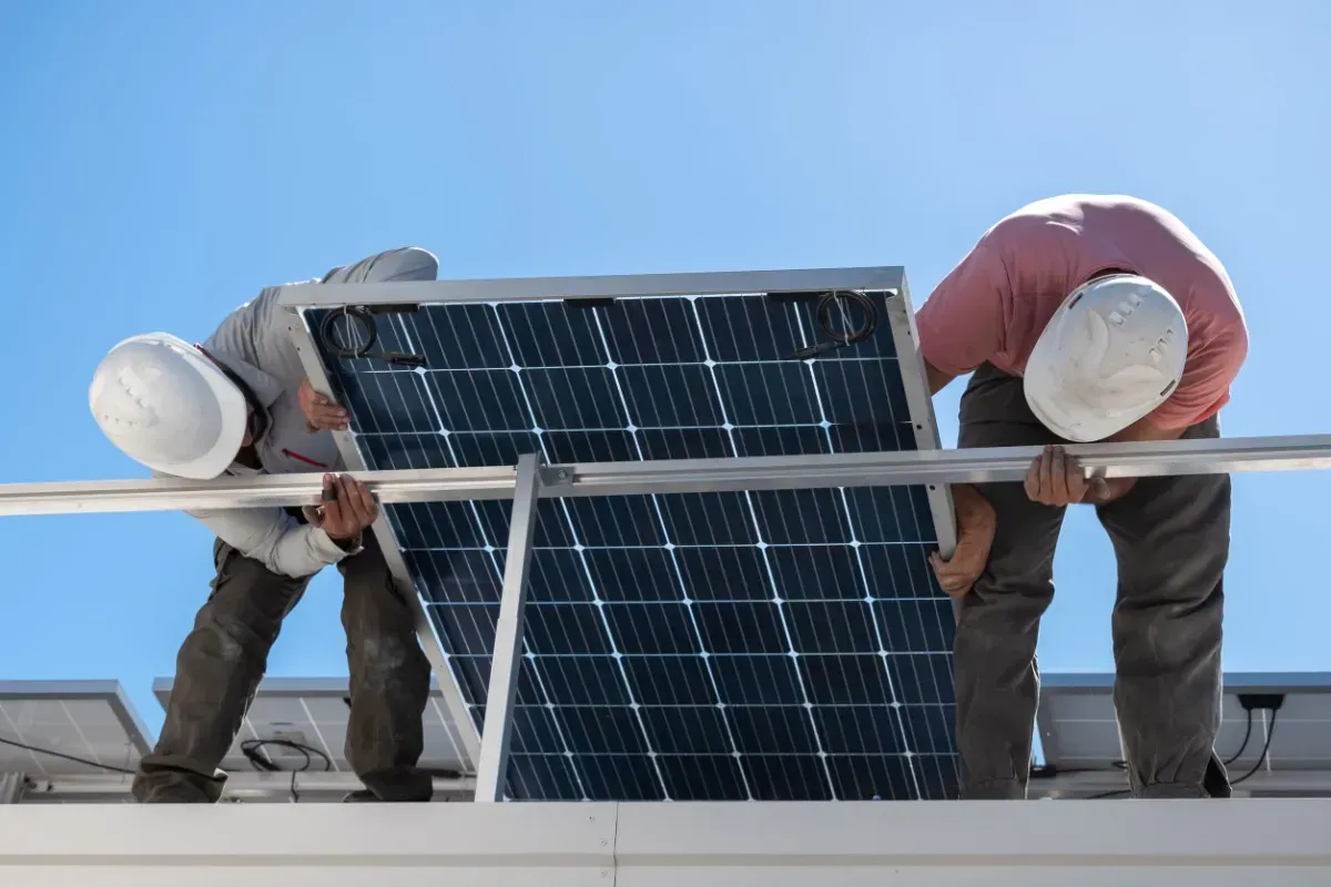 Dos trabajadores con casco instalando un panel solar en un tejado. Fondo de cielo azul.