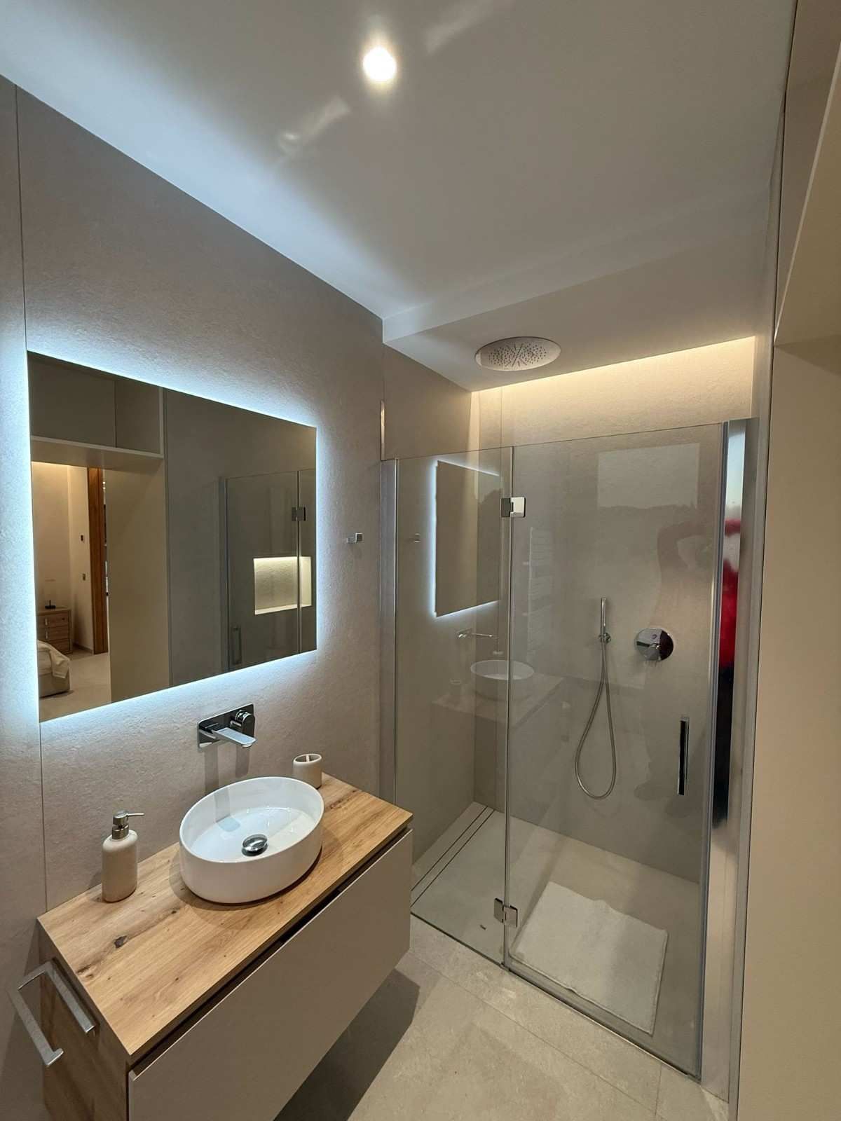 Un baño moderno con un lavabo blanco sobre un tocador de madera, un espejo de tocador iluminado y una ducha con mampara de vidrio.