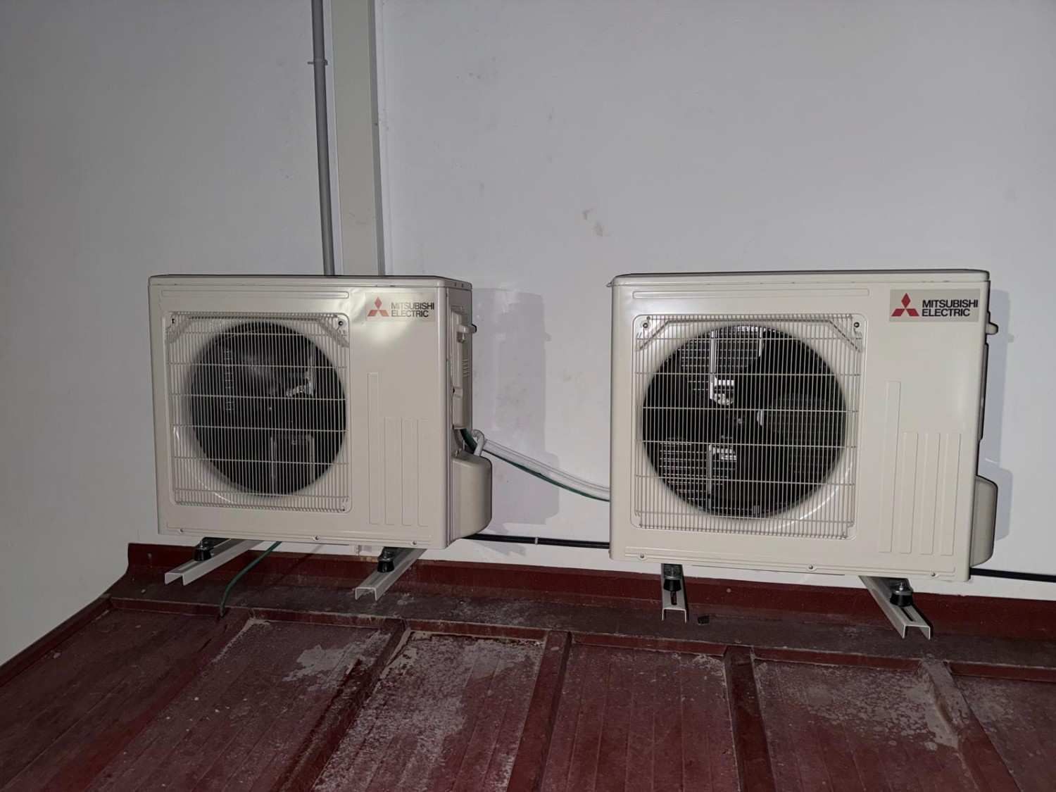 Dos unidades exteriores de aire acondicionado Mitsubishi montadas una al lado de la otra sobre un techo oscuro y corrugado contra una pared blanca.
