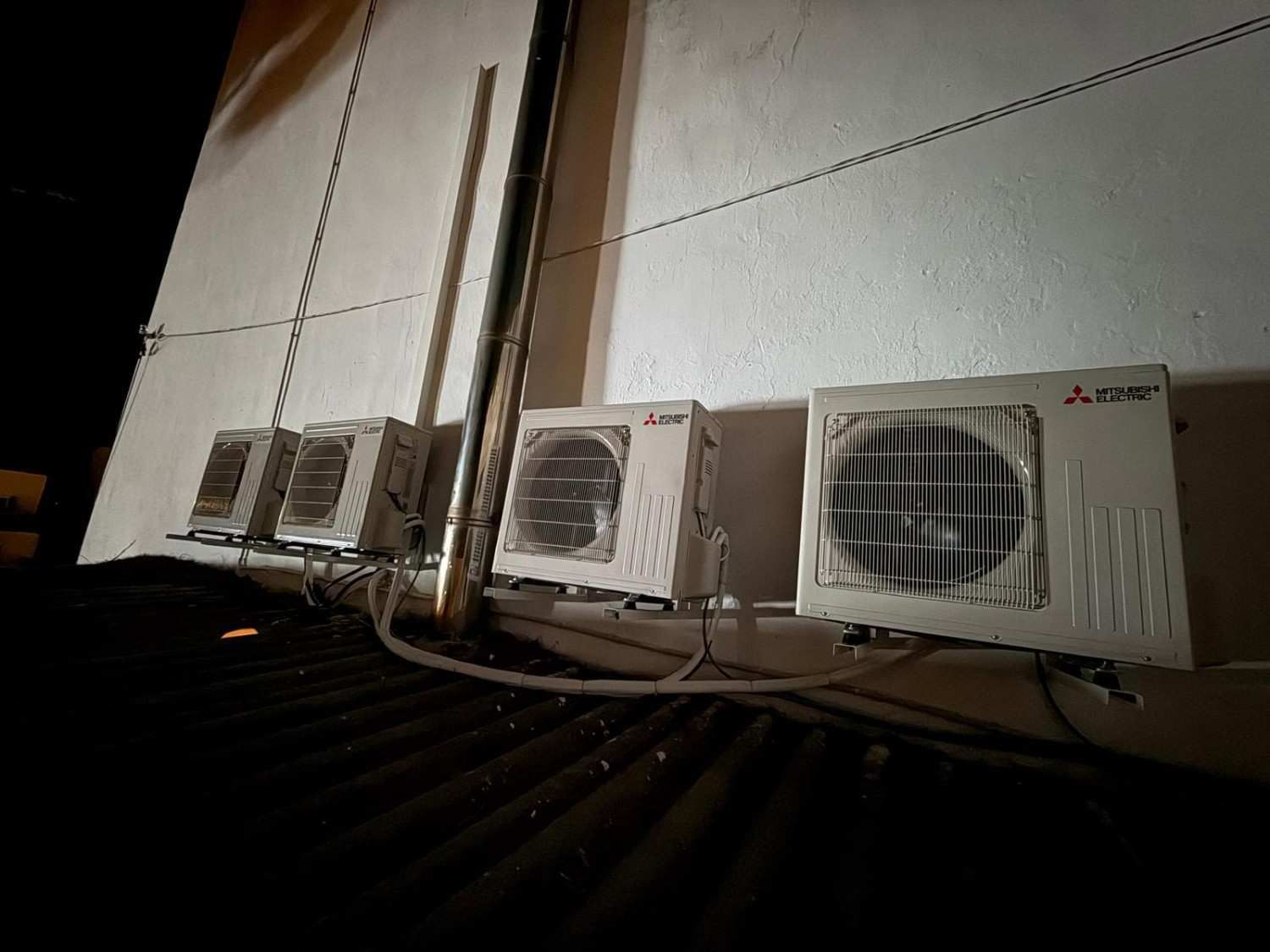 Cuatro unidades de aire acondicionado exterior Mitsubishi montadas en una repisa contra una pared exterior blanca por la noche.