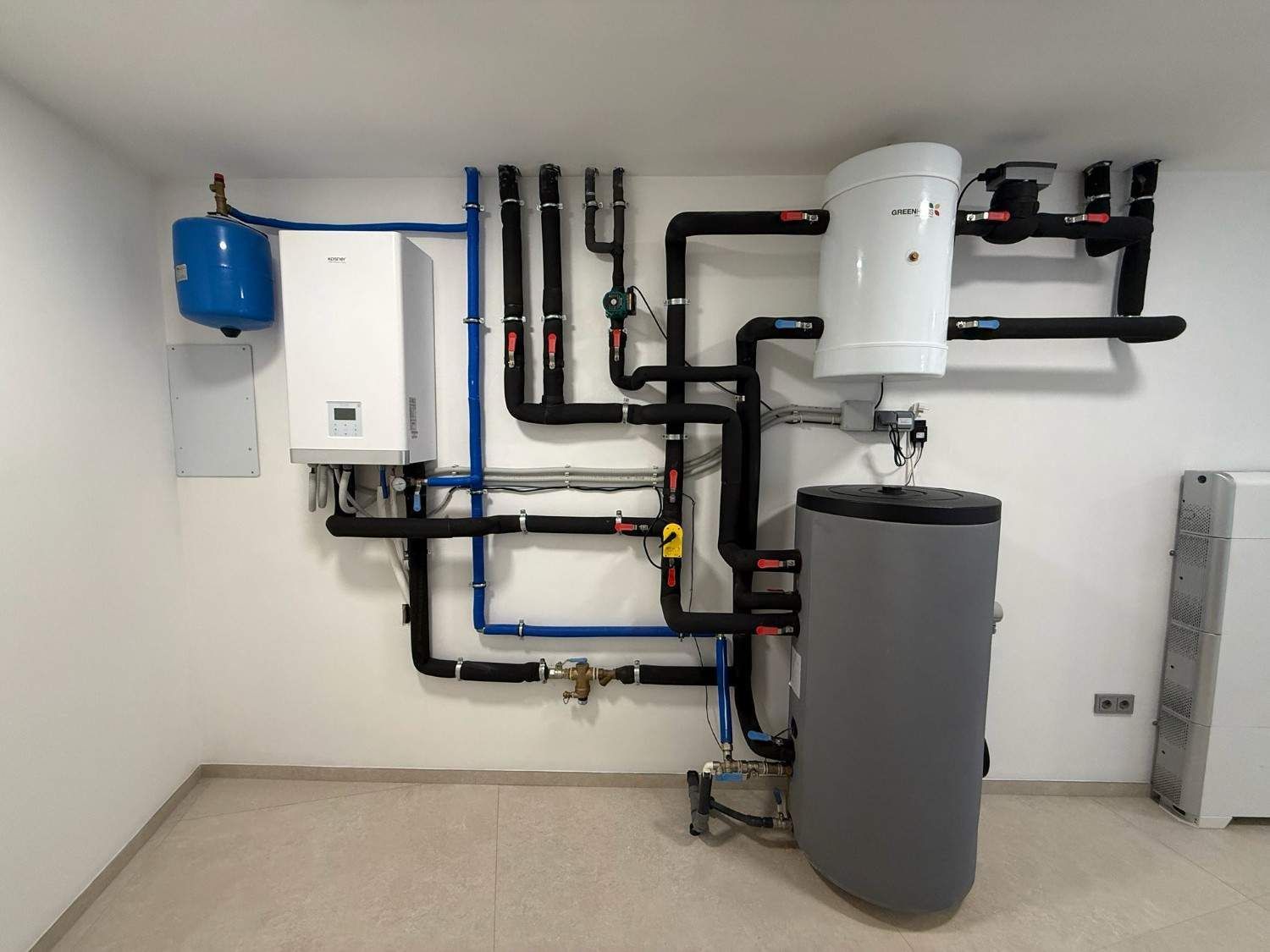 Un sistema de caldera y tanque de agua montado en la pared con tuberías negras interconectadas y un tanque de expansión azul en una habitación blanca.