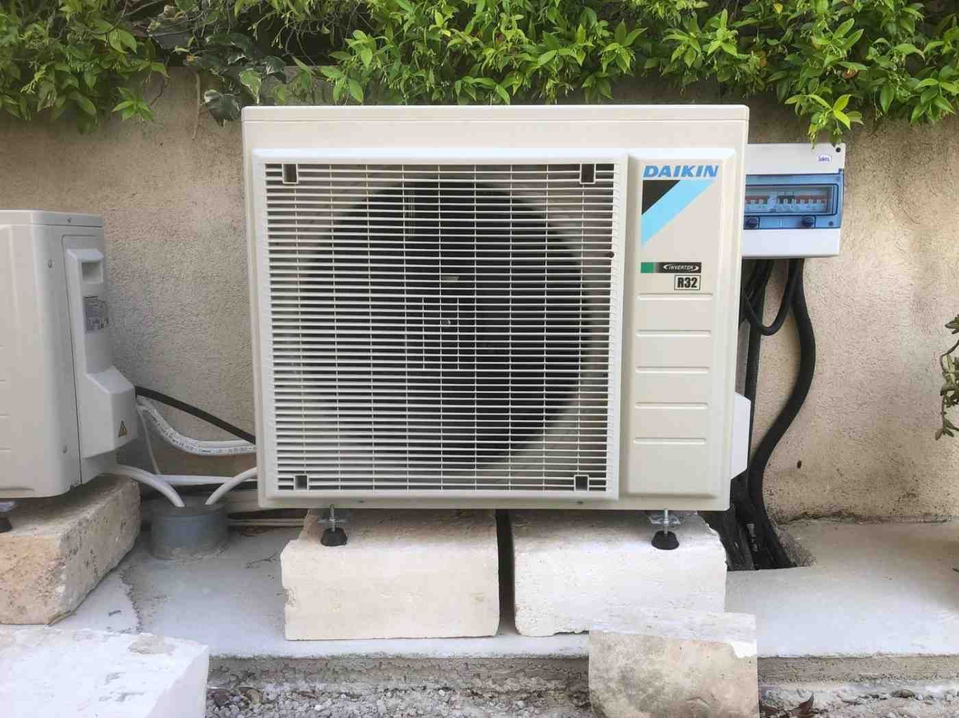 Una unidad exterior de aire acondicionado de la marca Daikin instalada sobre bloques de piedra contra una pared de color claro con follaje encima.
