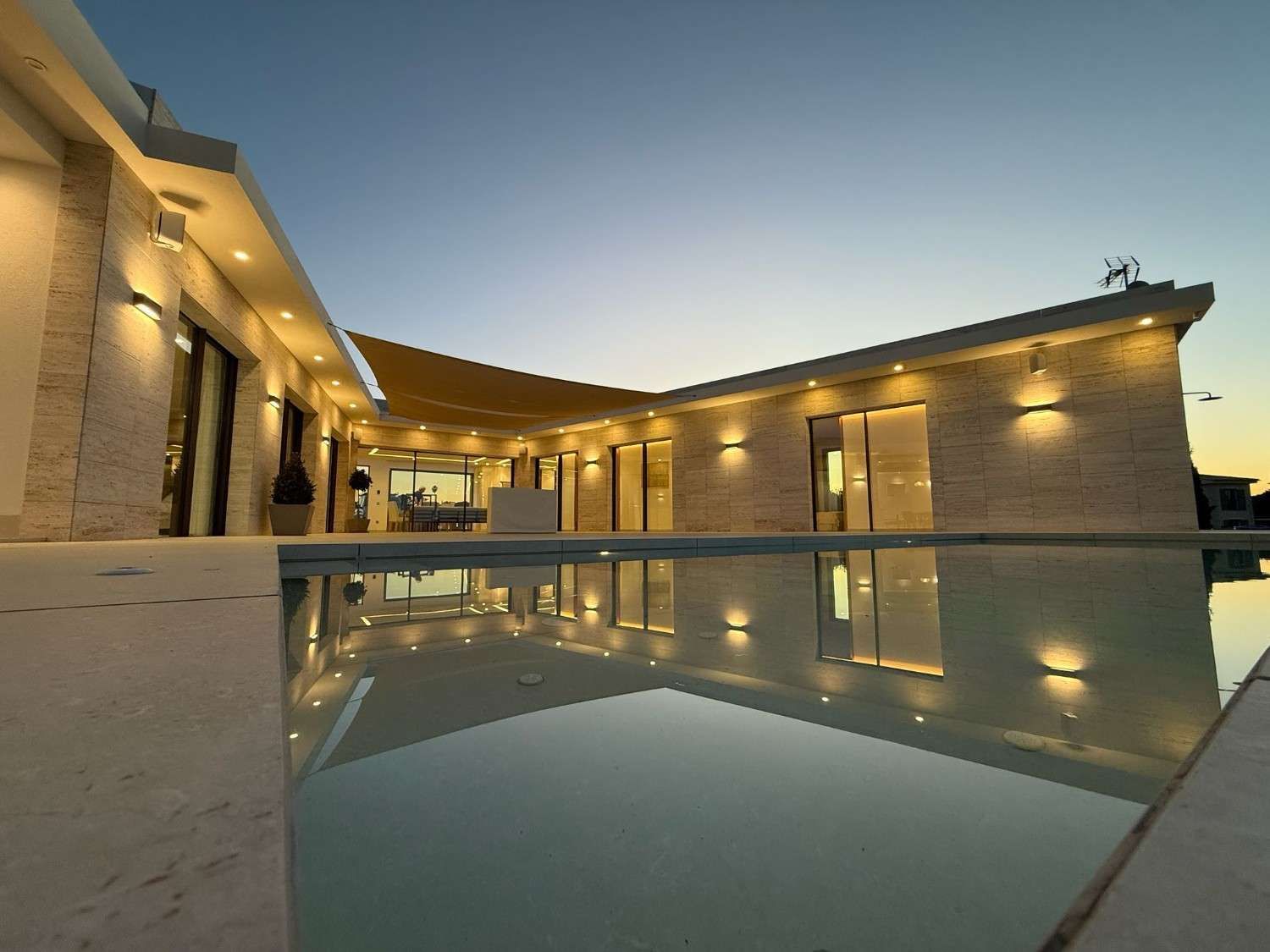Una vista de ángulo bajo de una casa de piedra moderna con paredes exteriores iluminadas al anochecer, reflejadas en una piscina tranquila.