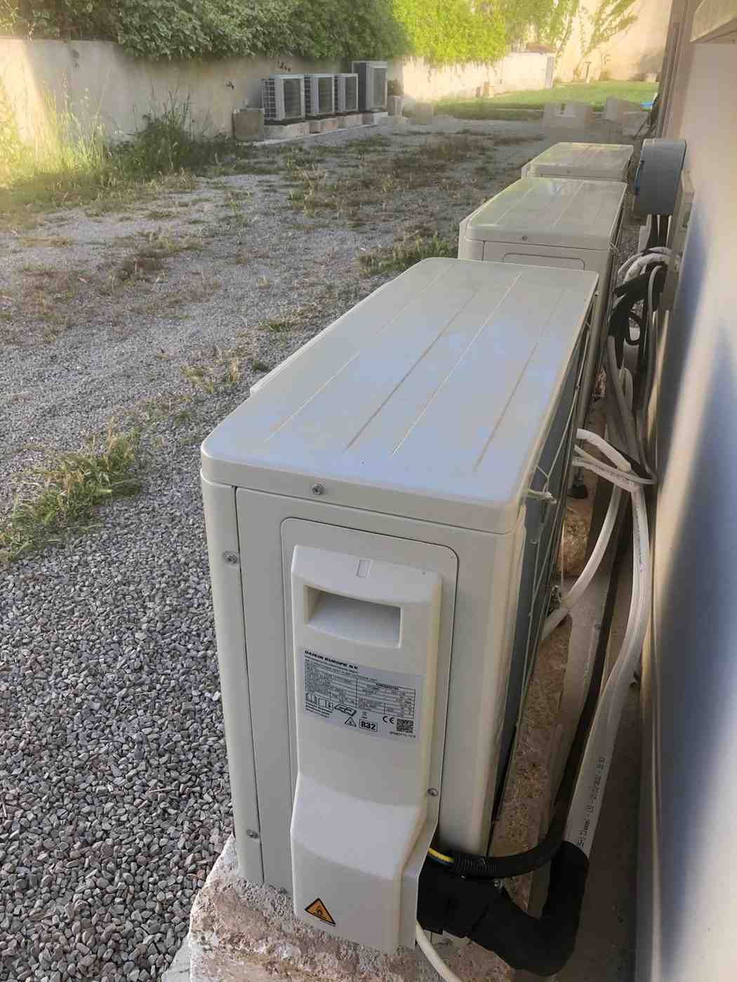Tres unidades exteriores de HVAC de color blanco se encuentran en fila sobre un suelo de grava al lado de una pared de edificio de color beige.
