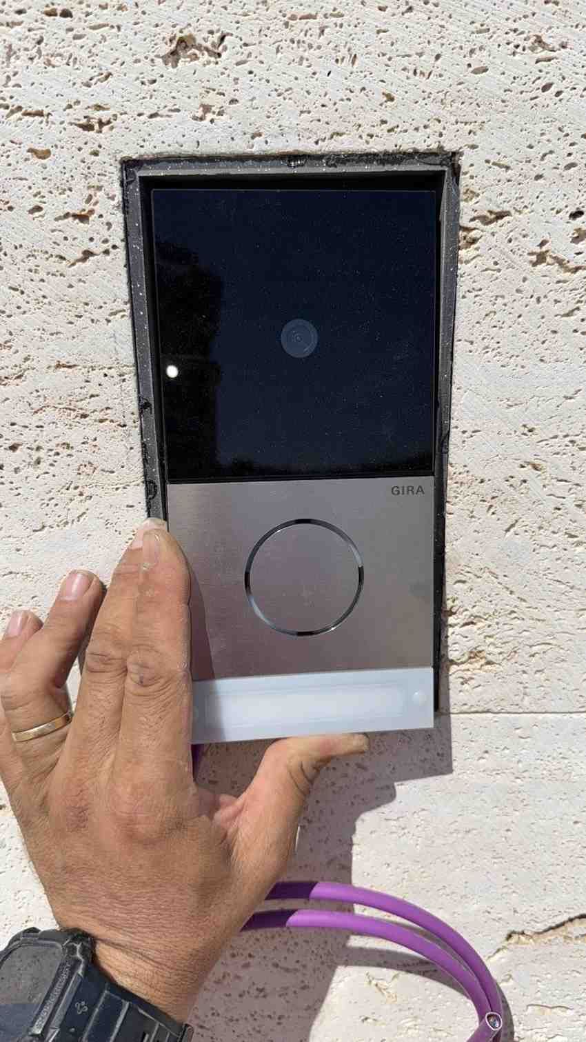 Una mano sostiene un timbre inteligente plateado y negro contra una pared de piedra blanca texturizada cerca de un cable violeta.