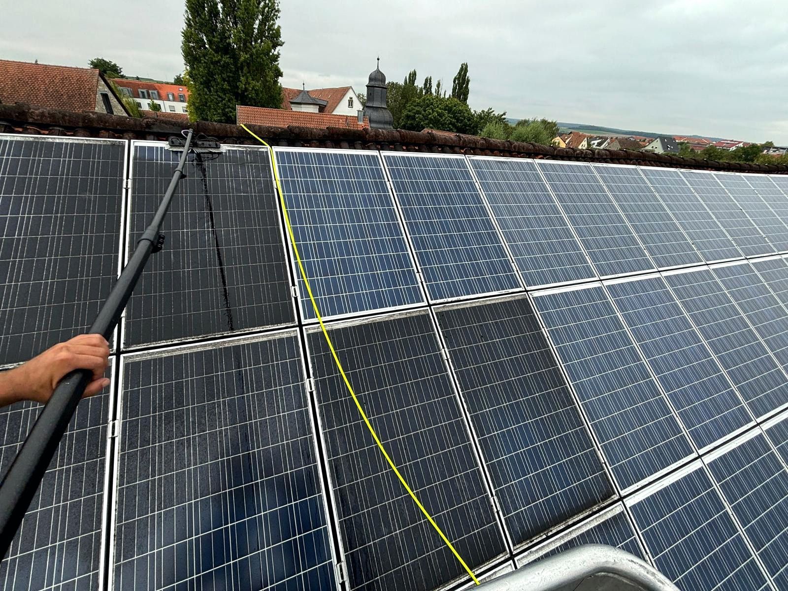 Eine Person reinigt mit einer langen Sprühlanze Solarpaneele auf einem Dach; die Paneele sind mit Wasser und Seife bedeckt.