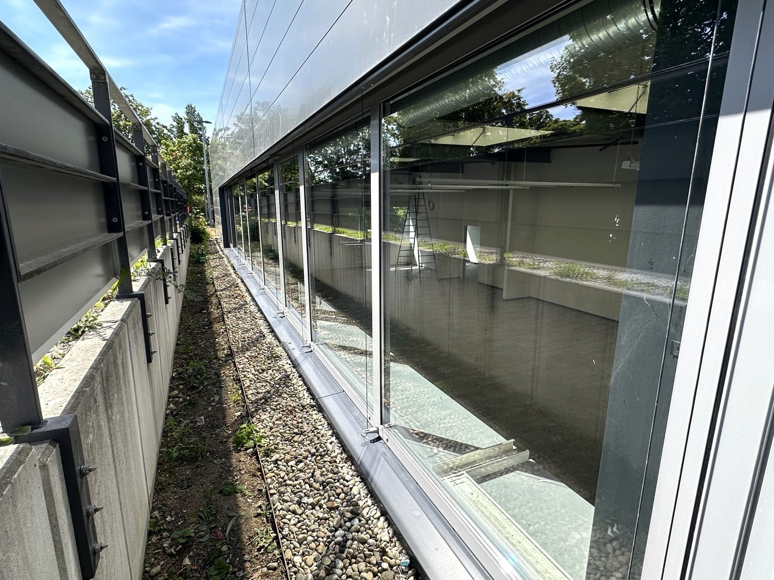 Ein schmaler Schotterweg zwischen einer Betonmauer und einem langen, spiegelnden Glasgebäude unter hellem Himmel