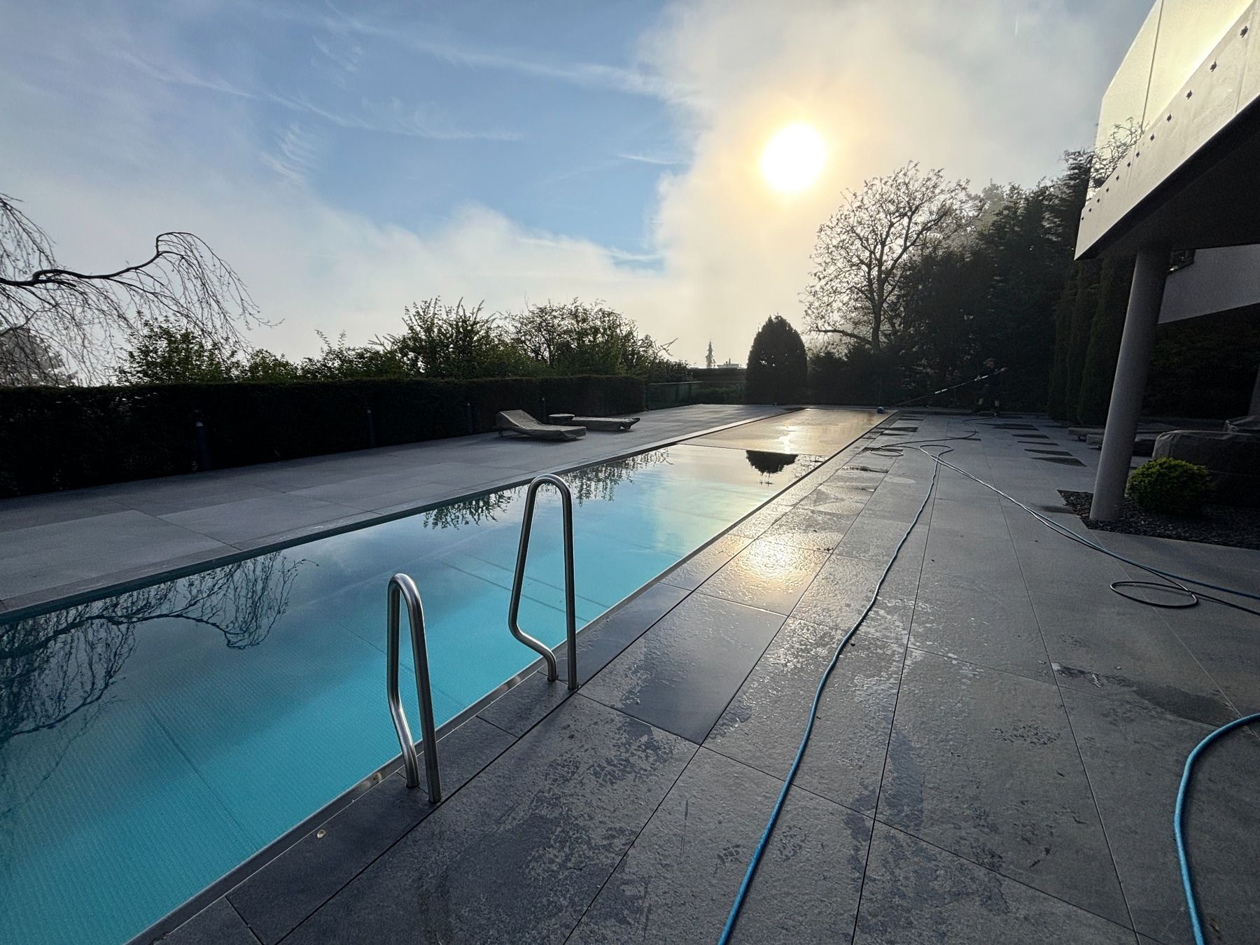 Außenpool bei Sonnenuntergang mit blauem Wasser, Leiter und nasser Terrasse neben Bäumen und einem Haus