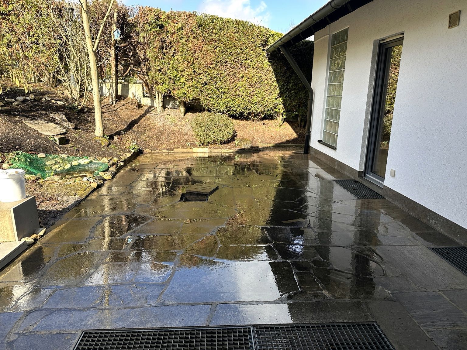 Eine feuchte Terrasse neben einem weißen Haus, in der sich der Himmel in Pfützen spiegelt, und ein von einer Hecke gesäumter Garten.