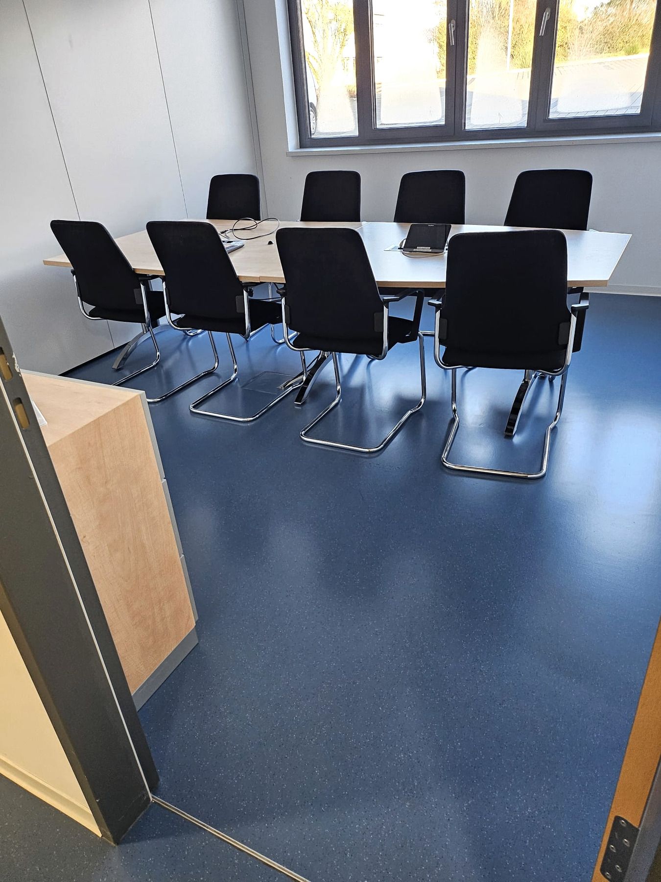 Konferenzraum mit weißem Tisch, schwarzen Stühlen, blauem Boden und großem Fenster.
